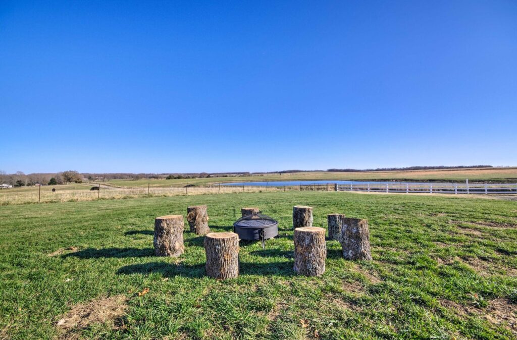 Outdoor Fire Pit – This Is My Temporary Home Fire pit surrounded by rustic tree stump seating on a grassy field, overlooking open farmland and a pond under a bright blue sky - This Is My Temporary Home.