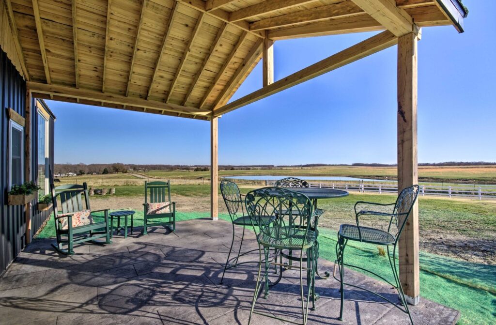 Covered Patio with Country View – This Is My Temporary Home Cozy covered patio featuring green metal chairs, a round table, and rocking chairs, overlooking wide open farmland and a distant pond under a clear blue sky - This Is My Temporary Home.