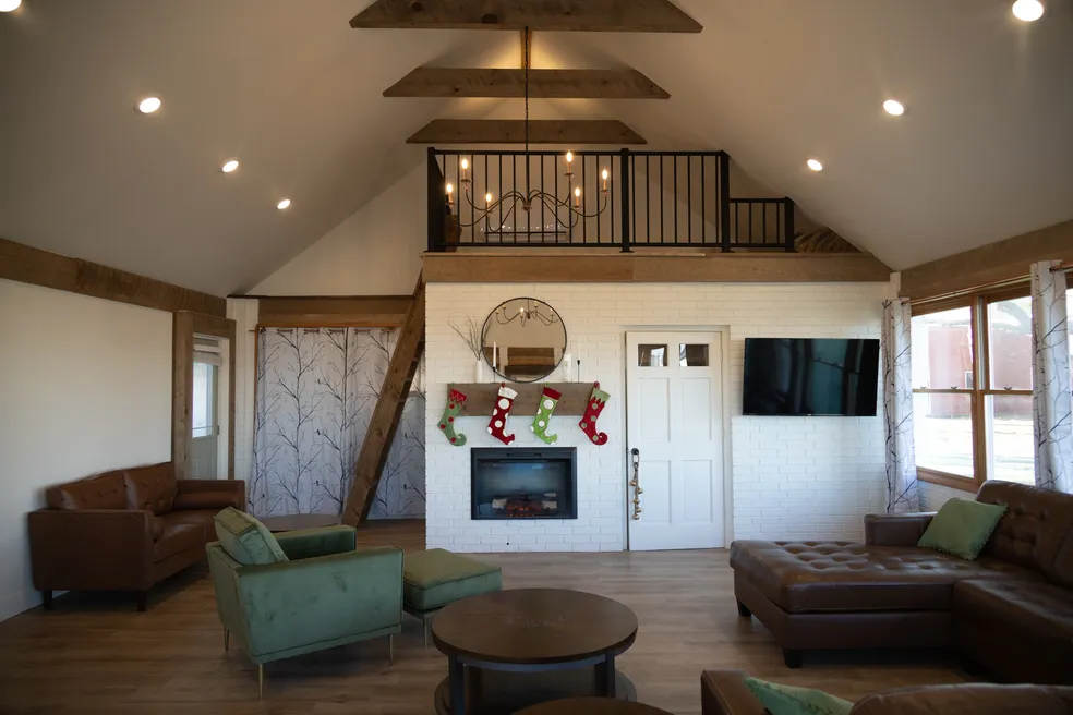 Cozy Living Room Interior – This Is My Temporary Home A cozy living room with vaulted ceilings, wooden beams, a loft with black railing, green and brown seating, a white brick fireplace decorated with Christmas stockings, and large windows – This Is My Temporary Home