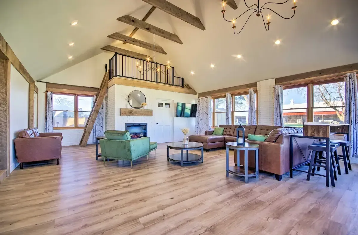 Spacious Living Room – This Is My Temporary Home Bright and spacious living room with vaulted ceilings, exposed wood beams, large windows, and cozy seating including green and brown chairs, at This Is My Temporary Home