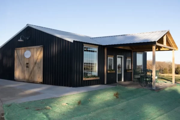 A black metal barn-style building with a light gray roof, wooden sliding door, and large windows, set against a clear blue sky – This Is My Temporary Home